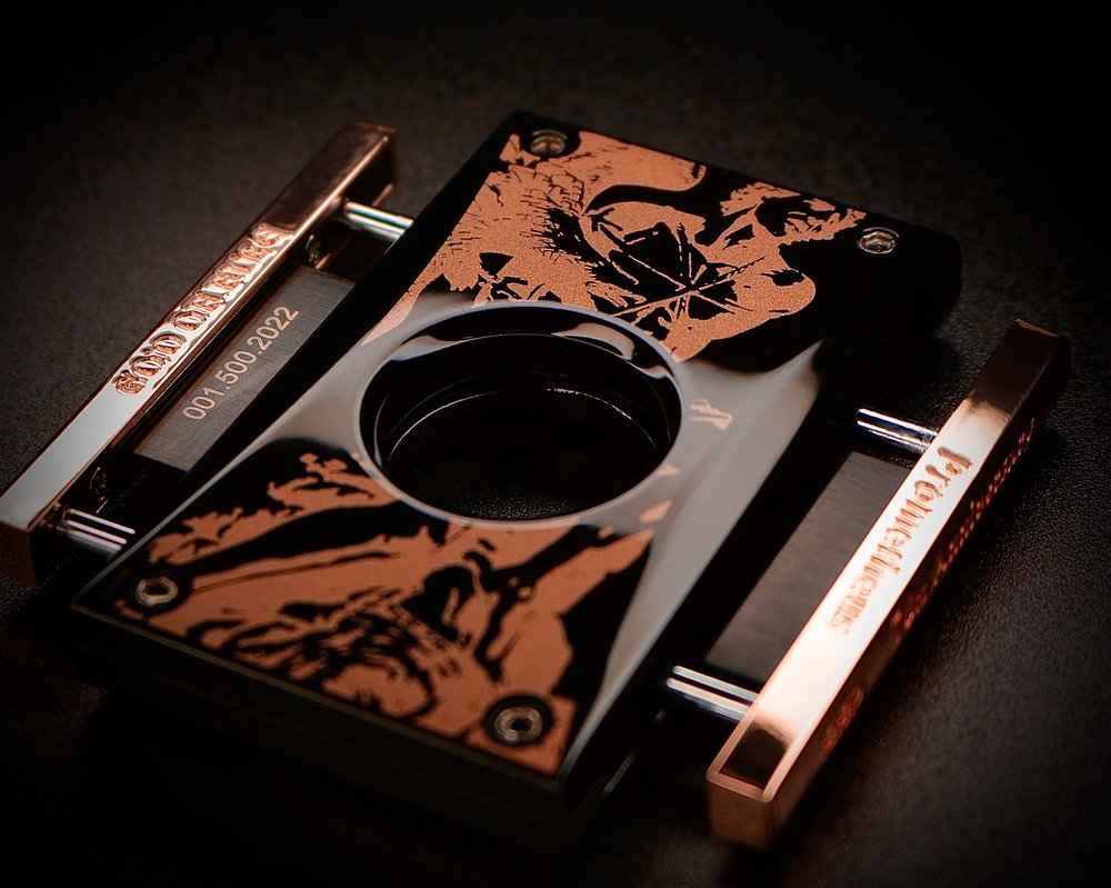 A vertical and sleek metal cigar cutter, black in colour with swirls of burnt umber accents. The cutter is from the Prometheus brand of accessories. The rose gold sides of the cutter come out and are squeezed to create the sleek and precise cut. The design is modern and good-looking. 