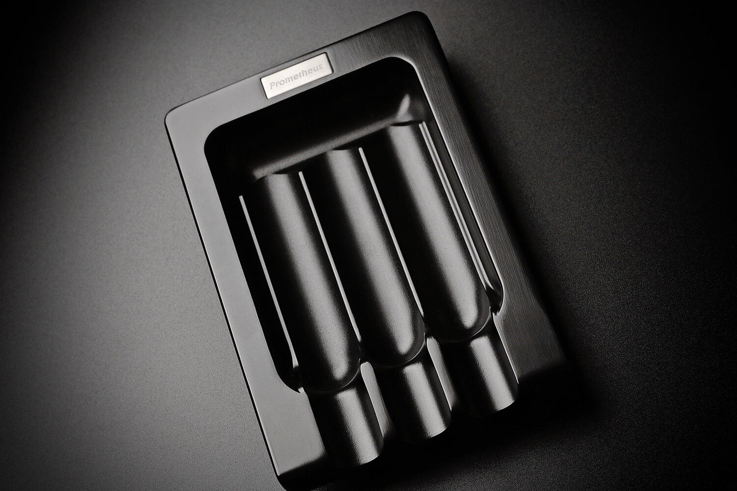 A modern black matte metal ashtray.  The ashtray is sleek with a triple cigar ledge. The brand is Prometheus.