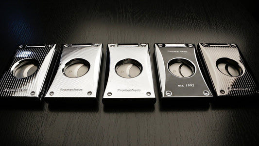 Five sleek and modern cigar cutters laid in a row. The cigar cutters are metal, each with a very classy design. The brand is Prometheus. 